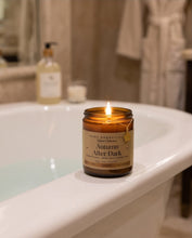 A lit amber glass jar candle labeled 'Autumn After Dark' by AOD ESSENTIALS, placed on the edge of a white bathtub in a softly blurred bathroom setting.