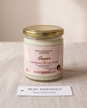 A glass jar candle with a gold lid and a pink and white label reading 'Signature Zodiac Collection Cancer The Heart of the Home' sits on a light fabric surface. A small white tag in front reads 'RCDC ESSENTIALS Bring comfort home.