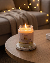 A lit candle in a clear glass jar sits on a round coaster on a wooden table, with a cozy living room background featuring a sofa and warm string lights.