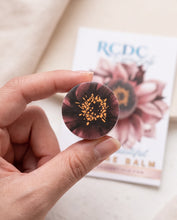A hand holds a small round balm puck with a dark burgundy and black floral pattern and gold stamen details, with an RCDC Beautiful Balm card in the background.