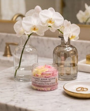 A jar of Sweet Honeysuckle whipped soap scrub sits on a marble bathroom counter, styled with white orchids in clear glass bottles and a small gold dish.