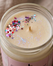 Top-down close-up of a candle in a clear glass jar with cream-colored wax, a single wick, and colorful round glitter confetti scattered across the surface. The jar has a pink label at the bottom and is photographed on a neutral beige surface.