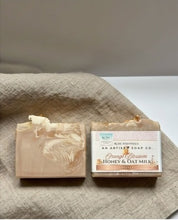 Two artisan soap bars displayed on a draped beige linen cloth. One bar is shown upright with its label facing forward, and the other lies on its side revealing a marbled swirl of cream, beige, and light brown.