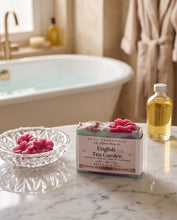 A bar of soap labeled 'English Tea Garden' by MCDC Essentials sits on a marble countertop in a bathroom setting, with a glass bowl of pink tea leaves, a bottle of oil, and a blurred bathtub in the background.