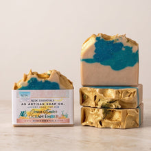 Three bars of handmade soap and one wrapped bar from Bed & Essentials An Artisan Soap Co. 'Ocean Amber' show their unique gold foil texture, with one bar sliced to reveal a blue and beige marbled center, set against a warm neutral background.