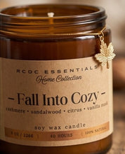 An amber glass jar candle with a kraft paper label reading 'RCDC ESSENTIALS Home Collection - Fall Into Cozy -' and a small gold maple leaf charm hanging from the rim.