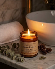 A lit amber glass jar candle labeled 'Something's Baking' by SCGC Essentials Home Collection, styled on a marble surface with eucalyptus leaves, pinecones, and a warm white vessel sink in the background.