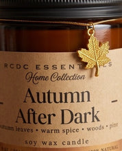 An amber glass soy wax candle jar with a kraft paper label reading 'RCDC ESSENTIALS Home Collection Autumn After Dark' and a hanging gold maple leaf charm.