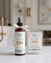 A bottle of room and linen spray and a scented melting wax melt in a clear clamshell package, both labeled RDCC Essentials and DW Hotel Lobby, set on a marble surface with a softly blurred bathroom background.