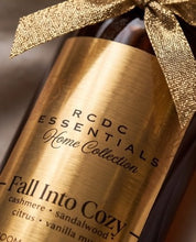 A close-up of a RCDC Essentials Home Collection 'Fall Into Cozy' candle bottle with a brushed gold label and a matching gold ribbon bow, set against a dark background.