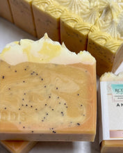 A close-up of a handmade soap bar with a marbled yellow and cream design, speckled with small black seeds, with other soap bars and a label partially visible in the background.