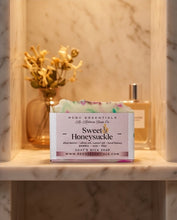 A boxed soap product labeled 'RCDC Essentials Sweet Honeysuckle' sits on a marble shelf in a warm indoor setting, with dried flowers and a bottle in the background.