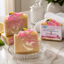 A image of handmade Pink Lemonade soap with pink and yellow colors, decorated with lemon slice designs, displayed with white towel and other decorative items.