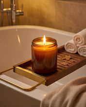 An amber glass jar candle with a lit flame sits on a wooden bath caddy draped over a white bathtub, with rolled towels and a warm bathroom setting in the background.
