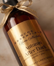A close-up of an amber glass bottle with a black cap and a gold ribbon bow, featuring a gold label that reads 'RCDC Essentials Home Collectibles Autumn After Dark' and 'Room + Linen Spray'.