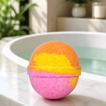 A spherical bath bomb with a pink base, yellow middle band, and orange top sits on the edge of a white marble tub with water and a softly blurred bathroom background.