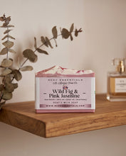 A bar of handmade soap labeled 'Wild Fig & Pink Jasmine' by RCOC Essentials sits on a wooden shelf. The soap has a pink and white marbled pattern, with a dusty pink paper label.
