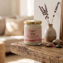A glass jar candle with a gold lid and a pink label reading 'Signature Scenic Collection Yogo The Grounding Effect' sits on a rustic wooden table beside a small vase with dried lavender and smooth stones in a softly lit room.
