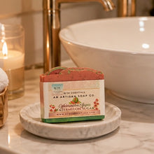 A bar of watermelon sugar soap by BCDC Essentials sits on a small marble dish on a bathroom counter, with a white vessel sink, gold faucet, and lit candle in the background.