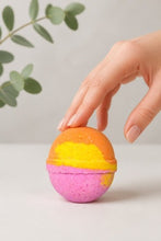 A hand with manicured nails gently touches a spherical bath bomb with a pastel sunset design of orange, yellow, and pink, set against a clean white background with soft eucalyptus leaves.