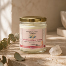 A glass jar candle with a gold metal lid and a pink label reading 'Signature Zodiac Collection Niro The Grounding Effect' sits on a stone surface with eucalyptus leaves and a clear crystal in a warm, softly lit setting.