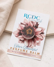 RCDC Essentials She's Beautiful Perfume Balm in a round tin, displayed on its illustrated floral backing card. The card features a pink flower design and is styled on a light beige textured surface with a folded cream fabric in the corner.