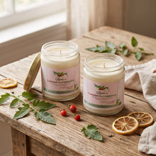 Two jars of Aesop's Garden Gatherings soy wax candles on a rustic wooden table, styled with green leaves, red berries, dried lemon slices, and a beige cloth near a window.