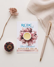 RCDC Essentials She's Beautiful Perfume Balm card with a small round balm tin placed on a cream surface, styled with a dried flower stem, a round floral object, and a pen.