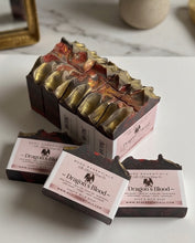 Three bars of handmade soap labeled 'Dragon's Blood' by Hode Essentials are arranged on a white marble-like surface. The center bar is standing upright in a dark wooden box with a pink label, while the other two lie in front.