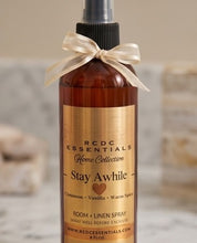 An amber glass room and linen spray bottle with a black spray top and a cream ribbon bow, labeled RCDC Essentials Home Collection Stay Awhile. The bottle is centered against a softly blurred beige and white background.