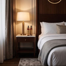 A styled bedroom interior with a neatly made bed, white bedding, a wooden nightstand, and a lit table lamp. A gold shopping bag icon with a dollar sign appears in the upper right against a dark wood wall.