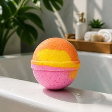 A spherical bath bomb with a pink bottom half, yellow middle band, and orange top half, resting on the edge of a white bathtub with a blurred bathroom background and green plant leaves.