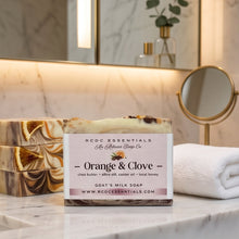 A bar of RPCD Essentials 'Orange & Clove' goat's milk soap is displayed on a white marble countertop in a bathroom setting, with a stack of similar soaps, a white towel, a small round mirror, and a glass bottle in the background.