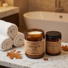 Two amber glass candles from SCBC Essentials on a marble bathroom counter, with rolled towels and autumn leaves nearby. One candle is open showing white wax, and the other has a black lid.