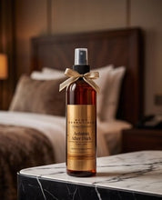 A bottle of MCOE Essentials 'Autumn After Dark' room spray, featuring an amber glass bottle, black spray nozzle, and a gold label with a beige ribbon bow, set on a marble-topped table in a softly blurred bedroom background.