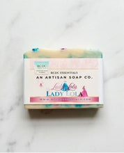 A rectangular bar of handmade soap with a white paper label reading 'Lady Lola' and 'RCDC Essentials' on a light marble surface. The soap has pastel green, cream, and blue marbled edges with small pink and blue decorative accents.