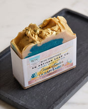 A single artisan soap bar in a white paper sleeve, photographed on a dark gray tray over a light marble surface. The soap has a cream base with a metallic gold top and a blue marbled section.