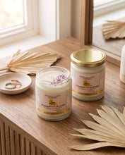 Two Mountain Living Collection scented candles, one open showing wax and dried flower petals, and one with a gold lid, styled on a wooden table with palm leaves and rings in a warm, natural setting.