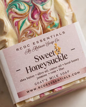 A close-up of a marbled soap bar wrapped in a paper label reading 'RCDC ESSENTIALS Sweet Honeysuckle' with visible cream, pink, teal, and gold swirls.