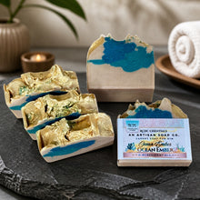 Handcrafted ocean-themed artisan soap bars with gold leaf and blue marbled details, displayed on a dark slate tray with a branded paper label in a styled spa-like setting.