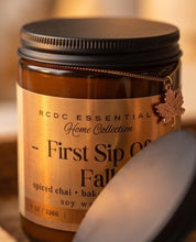 An amber glass jar candle with a black lid and a gold label reading 'RCDC ESSENTIALS Home Collection - First Sip Of Fall', shown with a blurred lid in the foreground against a warm, softly lit background.