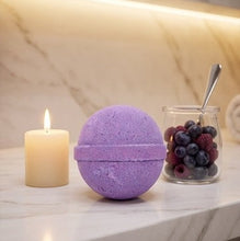 A purple spherical bath bomb sits on a marble countertop beside a lit cream candle and a small glass jar of berries with a spoon, against a softly lit marble backsplash.
