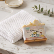 A bar of lemon poppy soap from Becca Essentials, showing the label and a small amount of the pale yellow soap, styled on a white waffle-knit towel over a white marble surface.
