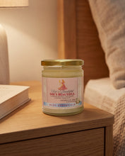 A glass jar candle with a gold lid sits on a wooden bedside table in a warmly lit bedroom. The label reads 'She's Beautiful' by BCPC Essentials.