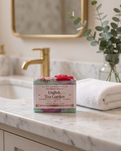 A rectangular bar of soap labeled 'English Tea Garden' by HooG Essentials sits on a white marble bathroom counter next to a folded white towel, with a blurred gold faucet and mirror in the background.