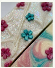 A close-up of decorative bars of soap, featuring one with a textured cream base covered in fine silver glitter and decorated with small teal and magenta flower shapes, and another bar showing a pink and teal marbled swirl pattern.