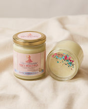 A glass jar candle with a gold lid and a pink label reading 'She's Beautiful' by BCDC Essentials, shown on a soft cream fabric background. One candle is upright and the other lies on its side to show the wax surface with colorful glitter.