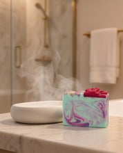 A rectangular bar of soap with a light blue and purple marbled pattern and a bright pink flower on top, placed on a marble bathroom counter next to a white dish with rising steam in a steam room.