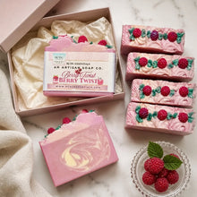 Artisan berry twist soap bars displayed in a pink gift box with cream tissue paper, alongside several loose soap bars decorated with pink swirls and red raspberry motifs, on a light marble surface with a small glass dish of fresh raspberries.
