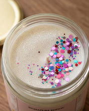 Top-down close-up of a candle in a clear glass jar with white wax and colorful round confetti on top, labeled 'SIGNATURE ZODIAC', on a wooden surface.